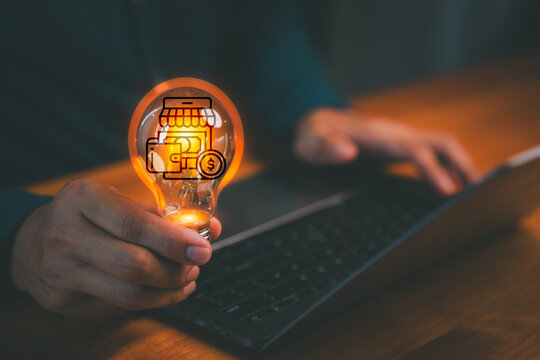 Hand holding glowing lightbulb with online store and wallet icons over laptop. Creative business idea, e-commerce innovation, startup success, and financial digital marketing concept.