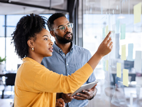 A woman and a man are collaboratively examining a glass wall covered with yellow sticky notes in a modern office.