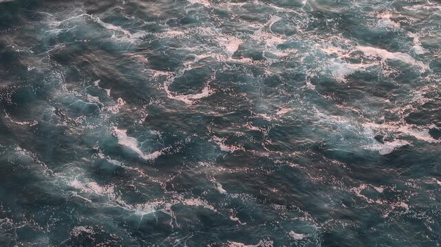 Aerial top-down view of turbulent ocean water with foamy waves and soft pink sunset reflections. Moody teal tones, dynamic motion and textured patterns evoke energy, nature and coastal atmosphere
