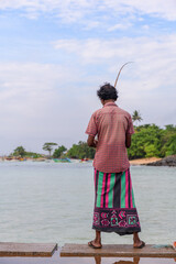 Fototapeta premium A traditional fisherman stands on a coastal pier holding a long fishing rod, facing calm tropical waters and a lush shoreline