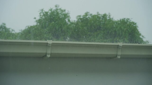 Heavy rain falling on house roof with green trees background during tropical rainy season.