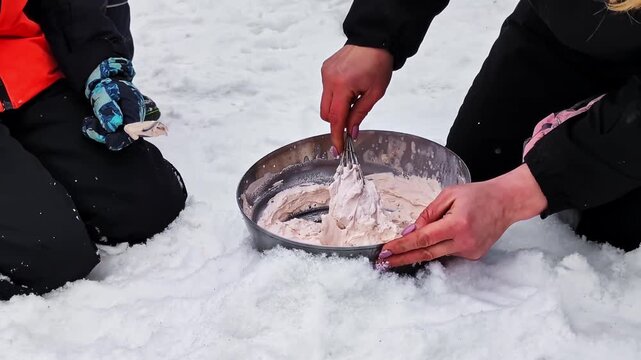 Close-up of female hands whisking a mixture in a metal bowl on the snow with a child watching