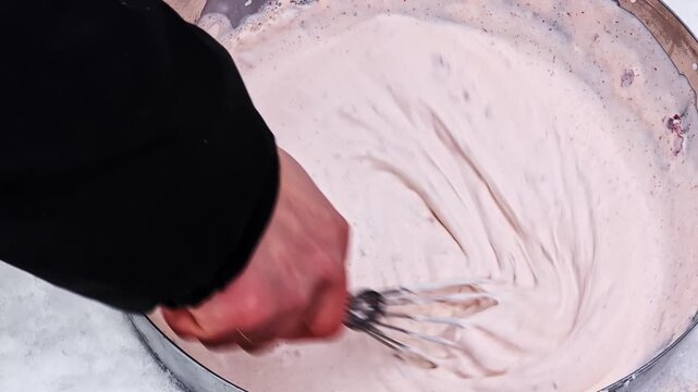 Close-up of a pastry chef's hands whisking a pink creamy mixture in a large metal bowl outside in snow