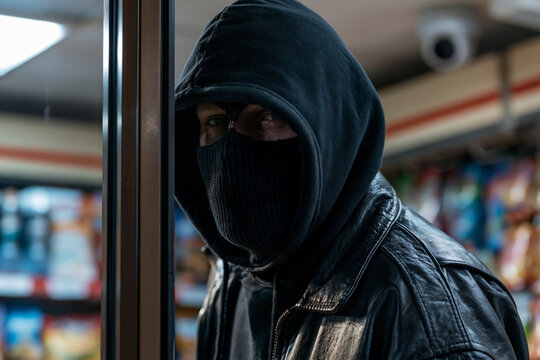 Dark hooded figure peers through store doorway. Face obscured, eyes watch with intense focus. Leather jacket adds gritty, stealthy atmosphere. Shelves and lighting blur behind the suspect