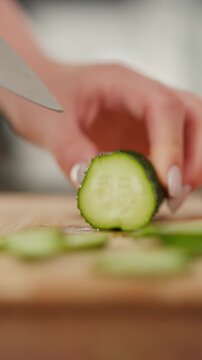 Hand slices cucumber into thin pieces on wooden cutting board focusing closely on action and texture details. scene emphasizes fresh produce preparation with soft lighting and shallow depth