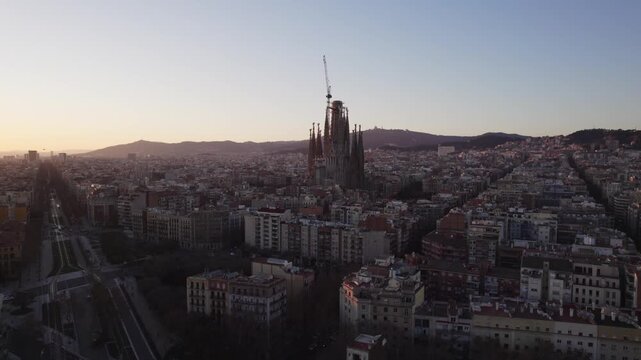 4K drone aerial view of Barcelona, Spain at sunset. The iconic Sagrada Fam&iacute;lia stands above the city&rsquo;s distinctive grid of streets, with the surrounding urban landscape and hills glowing in warm eveni