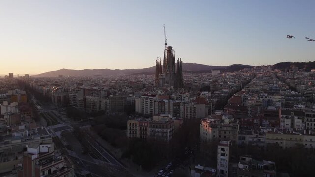 4K drone aerial view of Barcelona, Spain at sunset. The iconic Sagrada Fam&iacute;lia stands above the city&rsquo;s distinctive grid of streets, with the surrounding urban landscape and hills glowing in warm eveni