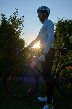 Cyclist in a helmet stands beside his bicycle outdoors at sunset