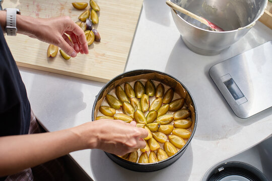 Woman arranging sliced plums on dough while making pie in kitchen.
