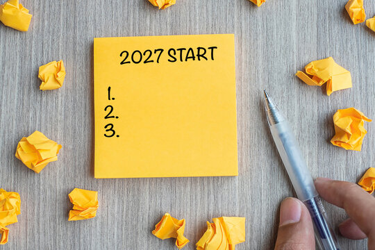 2027 Start word on yellow note with Businessman holding pen and crumbled paper on wooden table background. New Year, Resolutions, Strategy and Goal concept