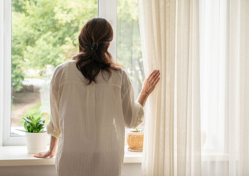 Middle-aged woman stands with her back to camera by window, parting light curtains and gazing at trees, immersed in morning reflection. Slow living, mindful lifestyle after 50
