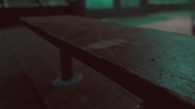 A worn wooden bench sits quietly, its surface marred by time and use. The soft glow of faded lights creates a mysterious atmosphere, hinting at stories left untold in this secluded spot.