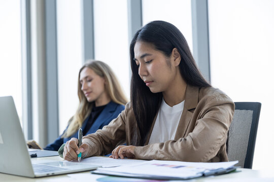asian business woman writing on document with laptop and caucasian coworker working together in corporate office environment