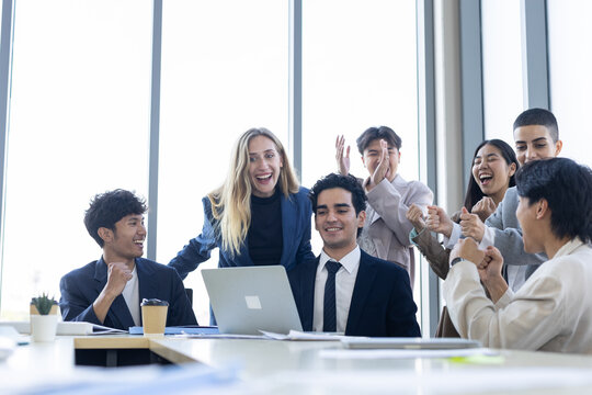 Joyful diverse business team celebrating success together around laptop in bright modern office, happy group feeling very excited