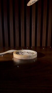 Close-up normal speed shot of whole coriander seeds being poured from a wooden spoon onto a small rustic wooden dish over a clean, shiny teak table.
