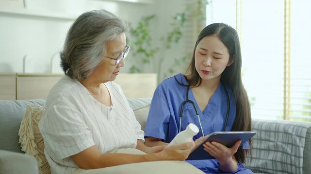 Professional Asian nurse explaining prescription medication to an elderly woman using a digital tablet during a home care visit. Reliable senior healthcare, medical assistance, and wellness concept.
