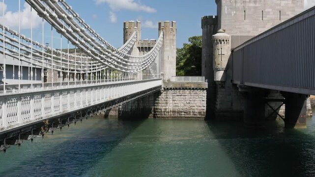 Conwy Suspension Bridge spanning river estuary beside Conwy Castle towers, stone masonry pier rising from green tidal water, North Wales
