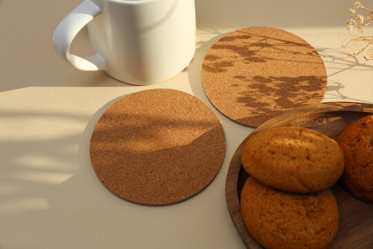 Cork round coasters and oatmeal cookies, ceramic cup on beige background with shadow from sunlight