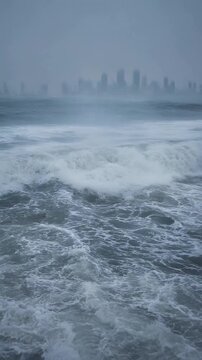Misty Ocean Waves Crashing Along Shore with City Skyline in Foggy Background