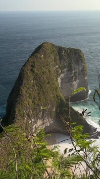 A rocky cliff rises above the ocean, covered in lush greenery. Waves crash against the shore below, creating a scenic coastal view.