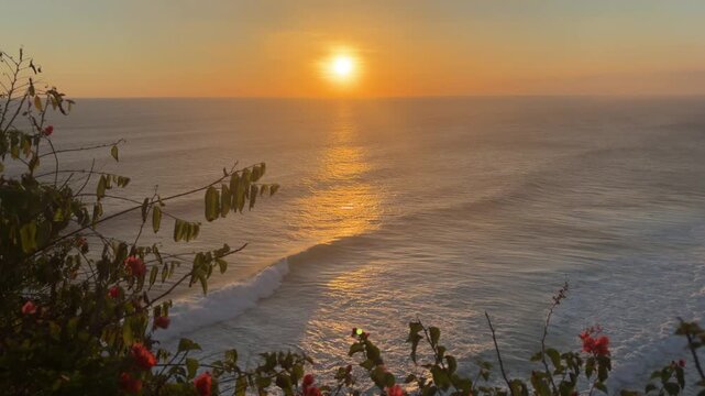 A serene sunset over the ocean, with gentle waves and vibrant colors. Silhouetted flowers in the foreground add a touch of nature to the tranquil scene.