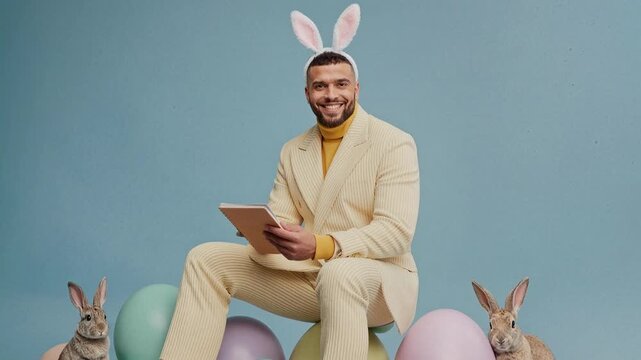 Man with bunny ears and rabbits celebrates Easter with pastel eggs. A smiling man wearing bunny ears and a stylish suit sits among pastel Easter eggs and rabbits, holding a tablet