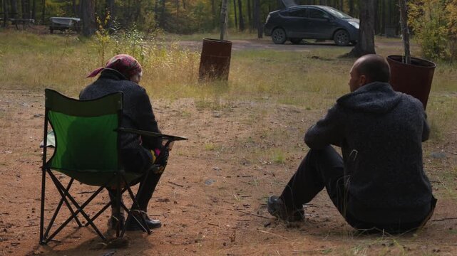 Two individuals converse while seated in a wooded area, one in a green folding chair and the other on the ground, with a parked car and rusted metal barrels visible behind them.