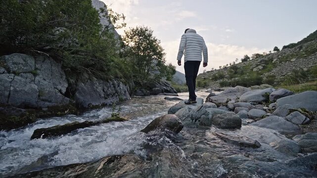 A man in a white jacket stands on a rock in a shallow mountain creek at early evening, surrounded by boulders and trees.