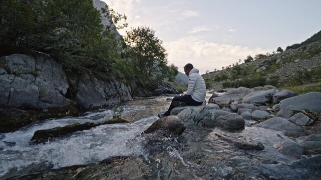 Man in a white jacket sits on a rock by a rushing mountain stream at dusk, relaxing in quiet alpine wilderness.