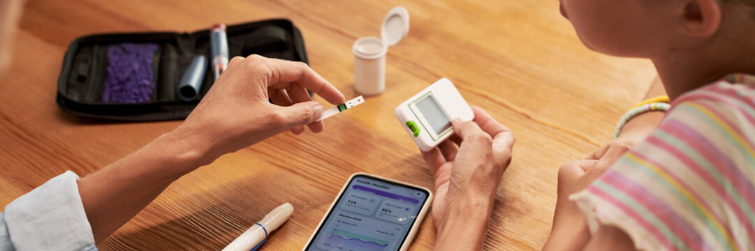 Close up of Caucasian adult helping Caucasian child test blood glucose using glucometer and smartphone app. Suitable for pediatric diabetes care and digital healthcare marketing