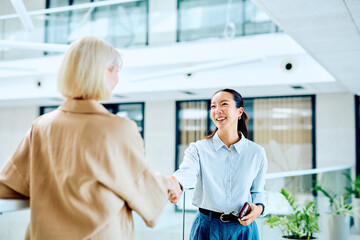 Two diverse businesswomen shaking hands, smiling and making an agreement in a bright modern office building. Professional collaboration © we.bond.creations