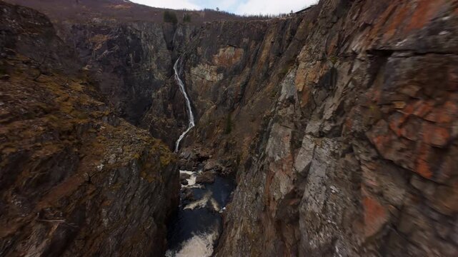 An FPV drone flies through a rocky gorge toward Voringfossen waterfall in Norway, revealing cascading water and steep canyon walls.