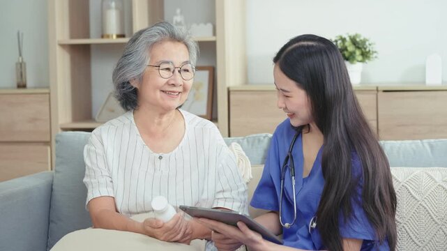Home care nurse reads prescription on tablet while elderly woman holds medicine bottle, going over dosage instructions on sofa in living room.