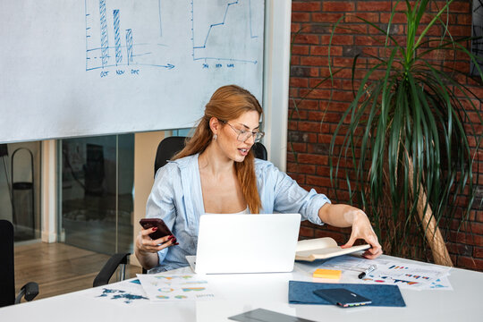 Business analysis meeting. Financial data report, Business and finance concept. Young woman sitting at the office desk, holding smartphone and turning the notebook page.