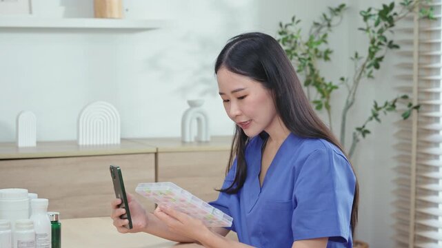 Side view of young nurse holding pill box up to smartphone, teaching patient how to sort daily pills during online telemedicine consultation.