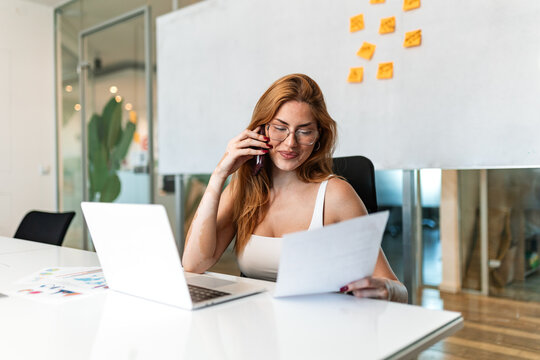 Business phone call. Corporate communication and task review, Office lifestyle concept. Young businesswoman sitting at the desk, talking on smartphone and reviewing financial report.