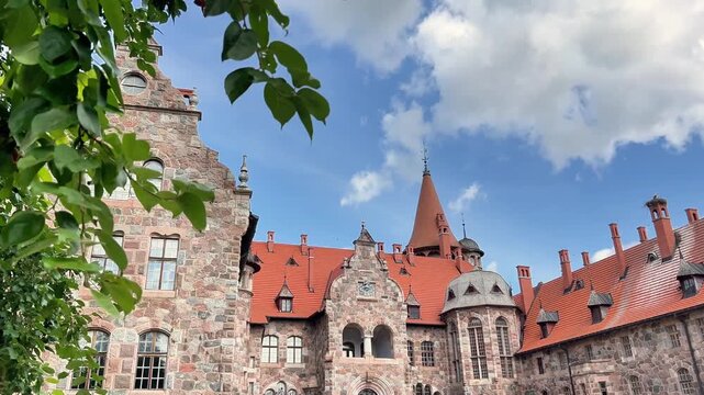 Exterior view of a historic stone castle building with a red tile roof and turrets framed by tree leaves. Ideal for travel documentaries, heritage tourism, and architectural media.