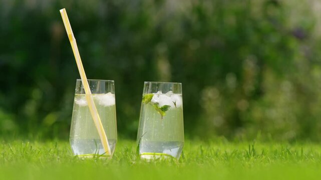 Two refreshing drinks with ice and mint decorated with cocktail umbrellas on grass.
