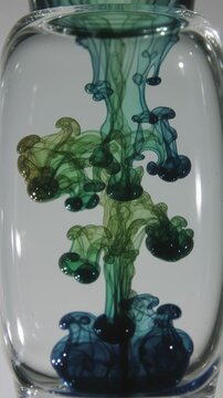 Colored ink drops diffuse through water inside a glass vessel, forming shifting plumes in an abstract fluid dynamics sequence