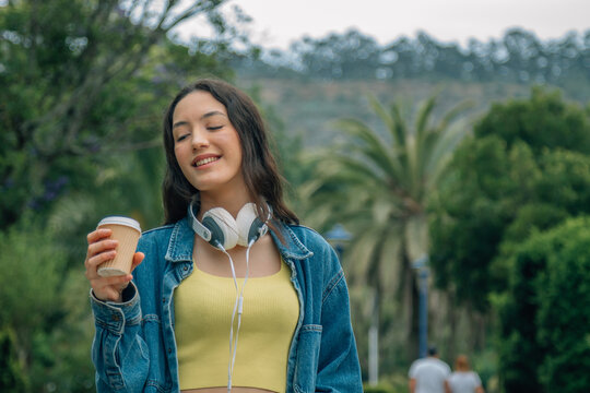 A relaxed young woman listening to music and drinking a takeaway beverage while walking down the street
