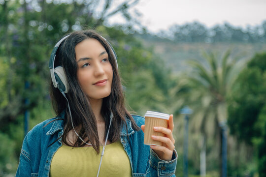 A relaxed young woman listening to music and drinking a takeaway beverage while walking down the street