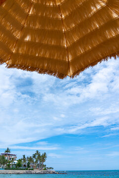 A Scenic View of a Tranquil Beach with a Thatched Umbrella Framing the Serene Sky and Ocean, Inviting Relaxation and Leisure in a Tropical Paradise