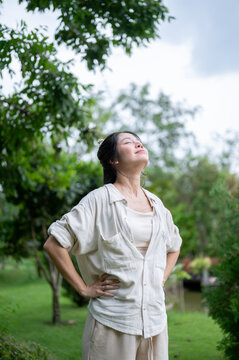 Asian woman standing with eyes closed and hands on hips in green outdoor garden.