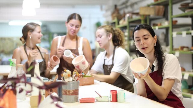 Interested young woman in pinafore drawing on handmade clay jar with paintbrush in art studio 
