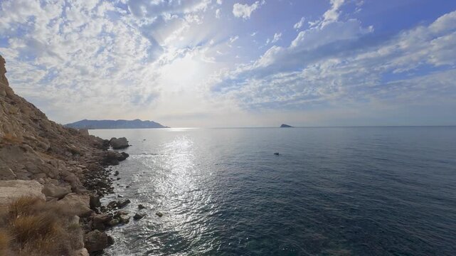 The view shows a coastline with rocky cliffs and the sea. Bright clouds cover the sky. The water reflects light. Some small boats are near the shore.