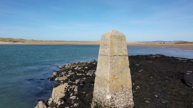 Aerial view of a stone trig point marker situated on the rocky shore of Ballyness Bay with sandy dunes and calm coastal waters in Gortahork, County Donegal, Ireland.