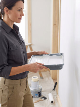Woman holding a paint tray while preparing to paint an unfinished interior. The scene represents home renovation, interior decoration and hands-on improvement work