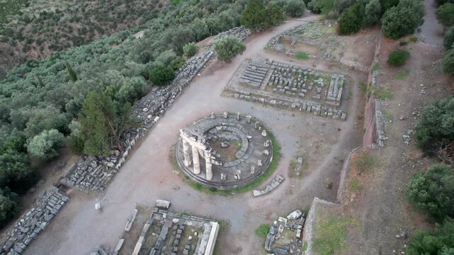 Aerial view of the ancient Tholos of Athena Pronaia ruins surrounded by olive trees and rocky terrain in the archaeological site of Delphi, Greece.