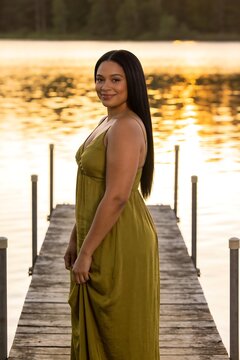 American woman in a sage-green dress standing on a lakeside dock at golden hour, peaceful lifestyle portrait with warm light, calm water, and natural elegance.