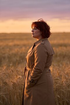 American woman in a camel trench coat standing in a golden wheat field at dusk, contemplative lifestyle portrait with warm amber light and a peaceful rural setting.